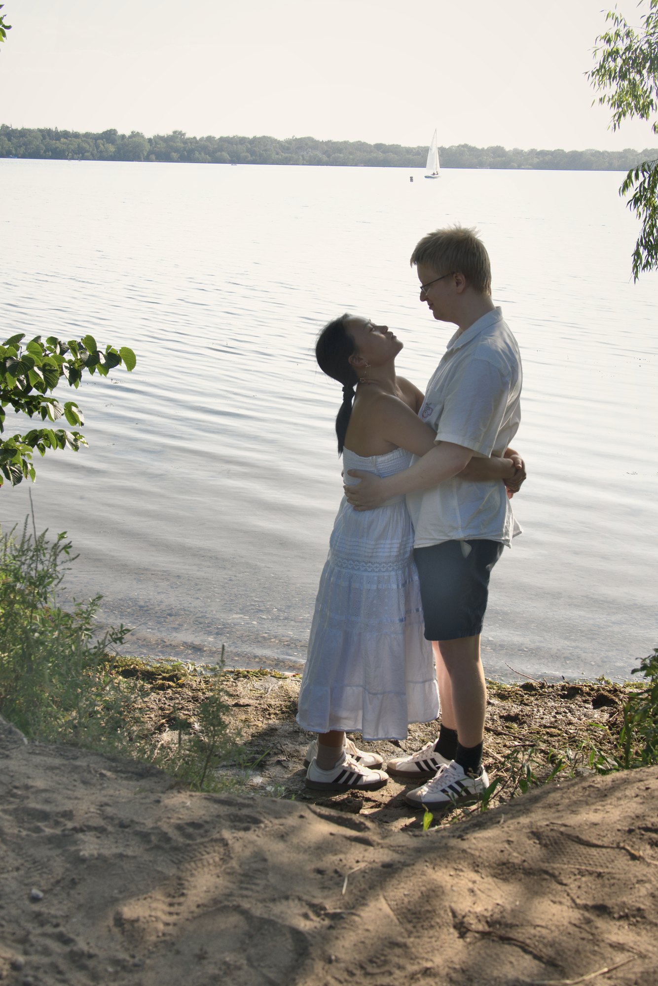 Couple embracing on a lakeshore with a sailboat on the water behind them — Twin Cities couples and engagement photography by Loon & Birch Photography